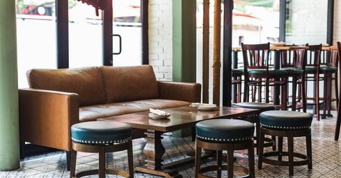 Cozy sunlit cafe interior with brown leather sofa, wooden coffee table, round upholstered stools on patterned tile floor and bar seating in the background