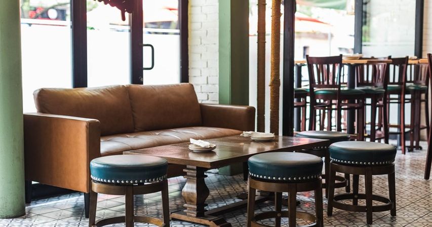 Cozy sunlit cafe interior with brown leather sofa, wooden coffee table, round upholstered stools on patterned tile floor and bar seating in the background
