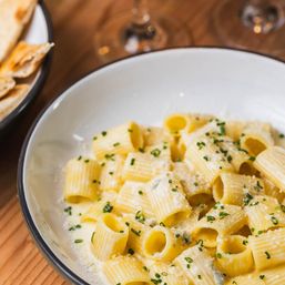 Creamy rigatoni pasta bathed in parmesan sauce, sprinkled with fresh chives, served in a white bowl on a rustic wooden table with warm flatbread at the side.
