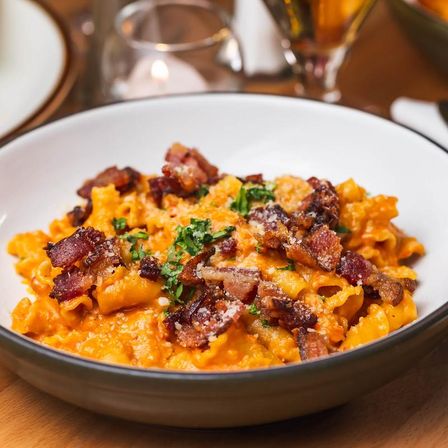 Creamy pumpkin pasta in a rich orange sauce topped with crispy bacon, grated Parmesan and chopped parsley, served in a white bowl on a wooden table in a cozy restaurant setting.