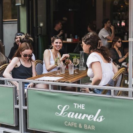Outdoor cafe patio with three friends chatting over cocktails and water at a wooden table with bistro chairs and green patio banner — sunny urban brunch vibe.