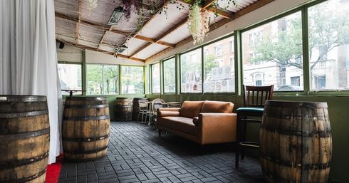 Cozy rustic enclosed patio lounge with wooden barrel tables, brown leather sofa, hanging plants and large windows overlooking a city street.