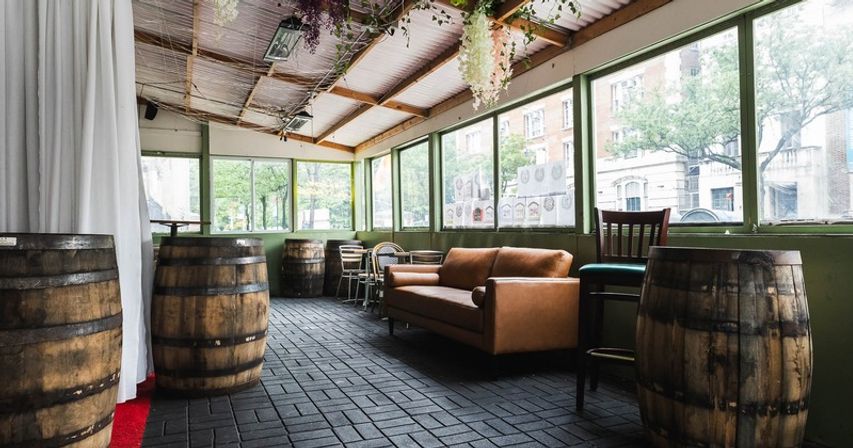 Cozy rustic enclosed patio lounge with wooden barrel tables, brown leather sofa, hanging plants and large windows overlooking a city street.