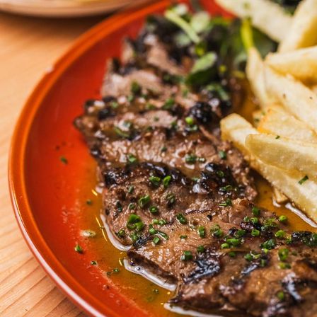 Close-up of sliced grilled steak bathed in savory jus, sprinkled with chopped chives and served with golden fries on an orange plate.