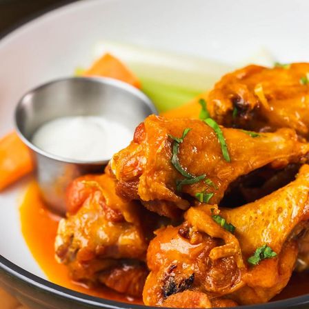 Crispy Buffalo-style chicken wings glazed in spicy orange sauce on a plate with creamy dipping sauce, carrot and celery sticks — classic game-day appetizer.