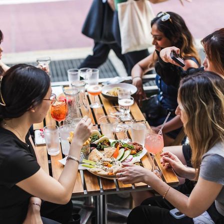 Friends dining at an urban sidewalk cafe patio, sharing Mediterranean mezze, pita, veggies and fries with colorful cocktails on a wooden table — one guest photographing the food.