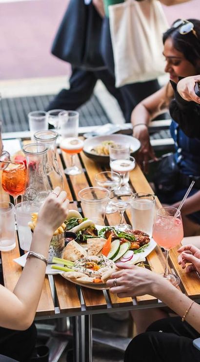 Friends dining at an urban sidewalk cafe patio, sharing Mediterranean mezze, pita, veggies and fries with colorful cocktails on a wooden table — one guest photographing the food.