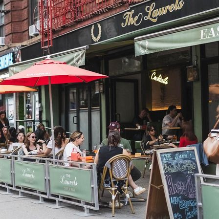 Outdoor city sidewalk café patio with red umbrellas and bistro chairs, groups of people enjoying drinks at tables, chalkboard happy-hour sign and neon "Cocktails" glow inside.