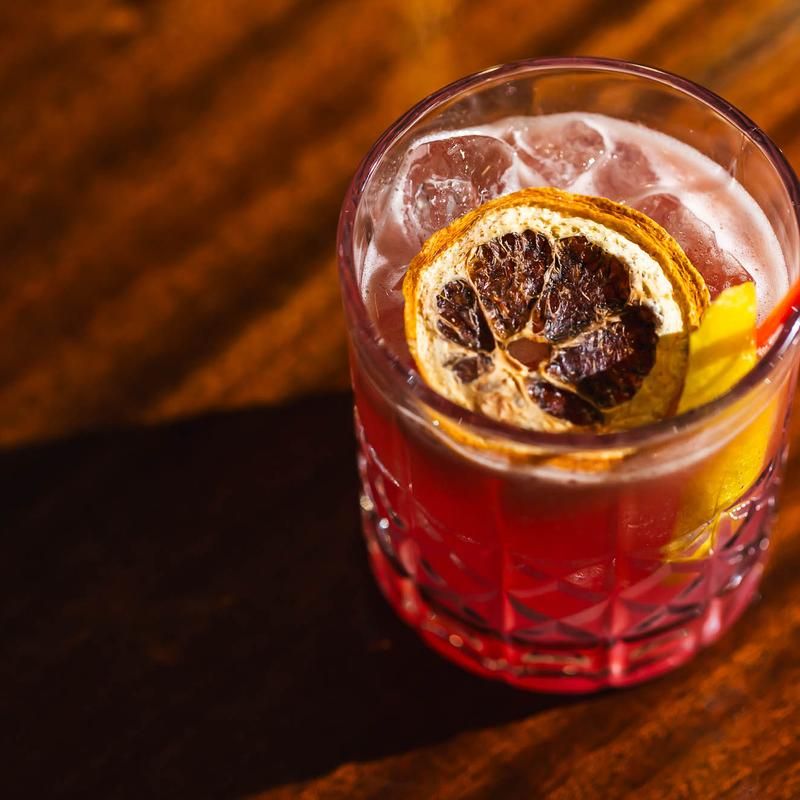 Vibrant red craft cocktail in a cut-glass tumbler with ice, a dried citrus wheel and lemon peel garnish resting on a wooden bar surface.