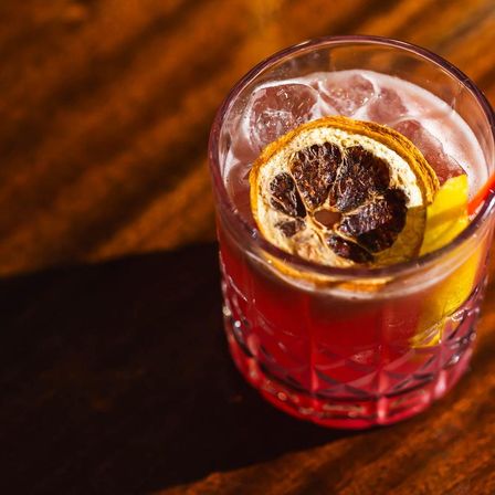 Vibrant red craft cocktail in a cut-glass tumbler with ice, a dried citrus wheel and lemon peel garnish resting on a wooden bar surface.