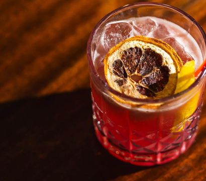 Vibrant red craft cocktail in a cut-glass tumbler with ice, a dried citrus wheel and lemon peel garnish resting on a wooden bar surface.