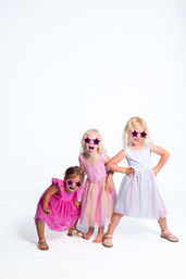 Three young girls in pastel party dresses and pink star-shaped sunglasses striking playful poses and laughing on a white studio background.