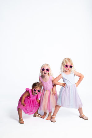 Three young girls in pastel party dresses and pink star-shaped sunglasses striking playful poses and laughing on a white studio background.