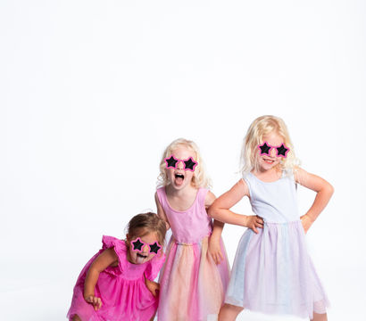 Three young girls in pastel party dresses and pink star-shaped sunglasses striking playful poses and laughing on a white studio background.