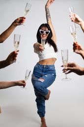 Woman in heart-shaped sunglasses, white crop top and ripped jeans strikes a playful pose holding a champagne flute during a champagne toast as friends raise flutes around her on a white studio background.