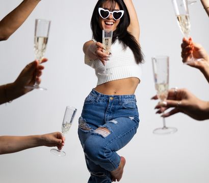 Woman in heart-shaped sunglasses, white crop top and ripped jeans strikes a playful pose holding a champagne flute during a champagne toast as friends raise flutes around her on a white studio background.
