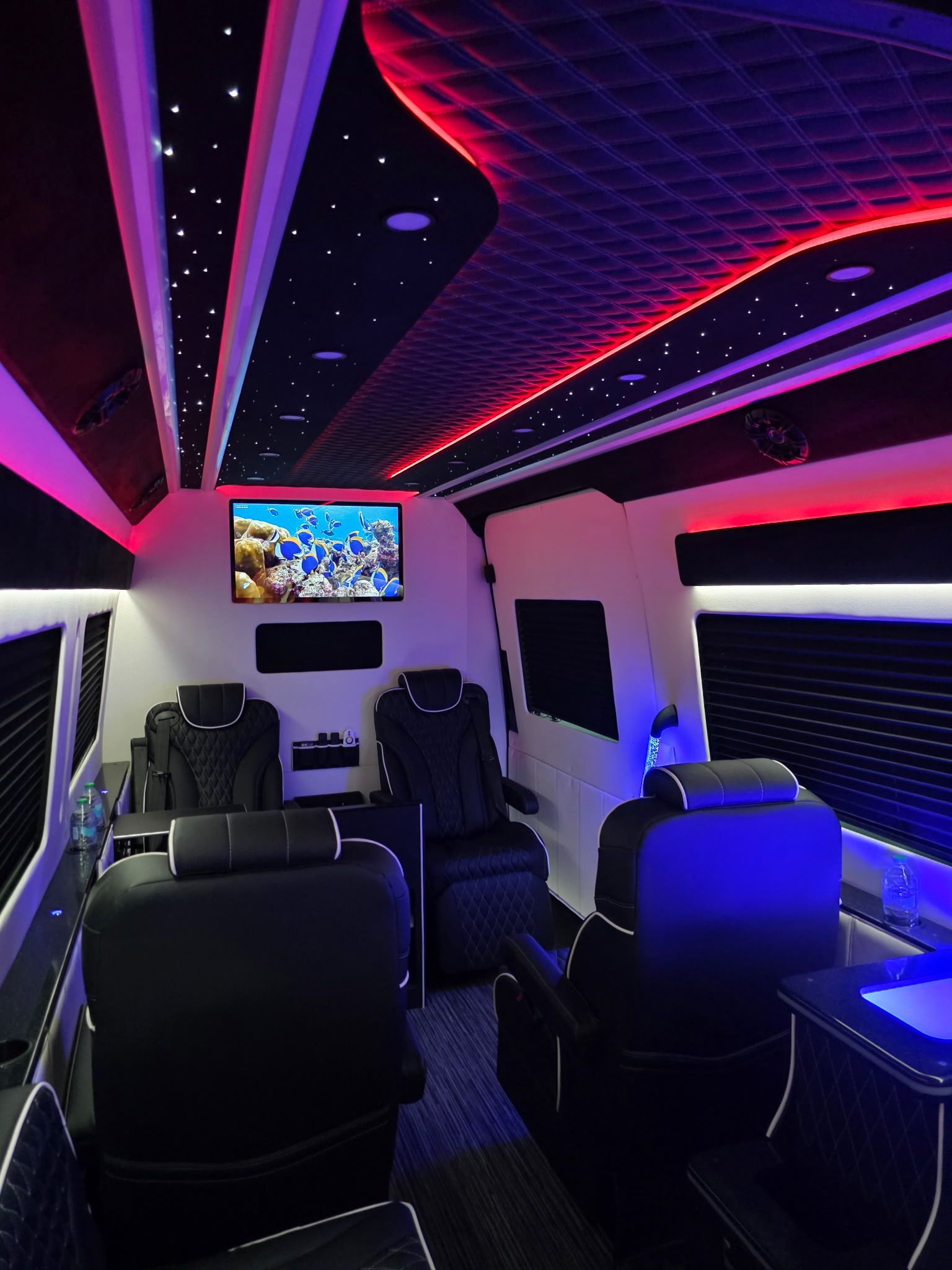 Luxury van interior with quilted black leather recliners, starry fiber-optic ceiling and red-blue LED accent lighting, wall-mounted TV showing a colorful coral reef — upscale mobile lounge.