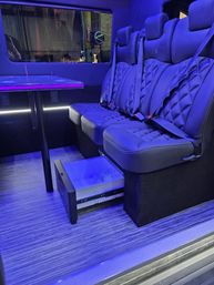 Sleek luxury van interior with quilted leather bench seats and seatbelts, a fold-down table, purple-blue LED ambient lighting and a pull-out mini fridge drawer over striped wood flooring.