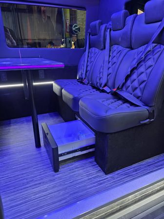 Sleek luxury van interior with quilted leather bench seats and seatbelts, a fold-down table, purple-blue LED ambient lighting and a pull-out mini fridge drawer over striped wood flooring.