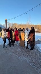 Group of eight women in colorful winter coats posing on a snowy patio under string lights, with bare trees and a clear blue winter sky.