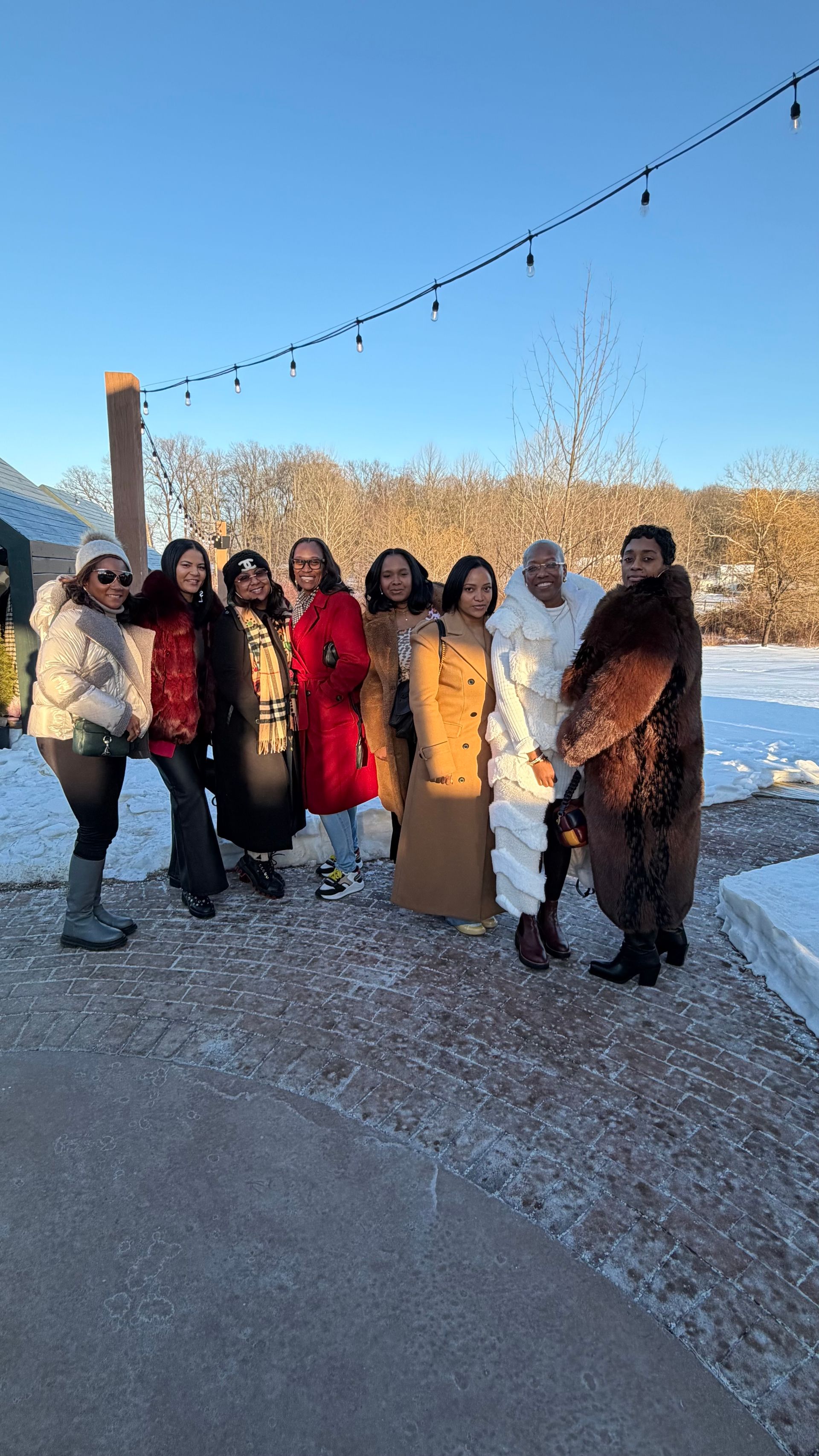 Group of eight women in colorful winter coats posing on a snowy patio under string lights, with bare trees and a clear blue winter sky.