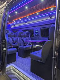 Luxury van interior with quilted black leather seats, folding table, built-in speakers and vibrant purple-blue and orange LED ambient ceiling lighting, modern VIP shuttle cabin.