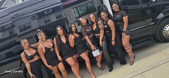 Eight women in coordinated black party outfits smiling and posing beside a glossy black party van on a suburban street, wearing heels, sunglasses and statement accessories for a night out.