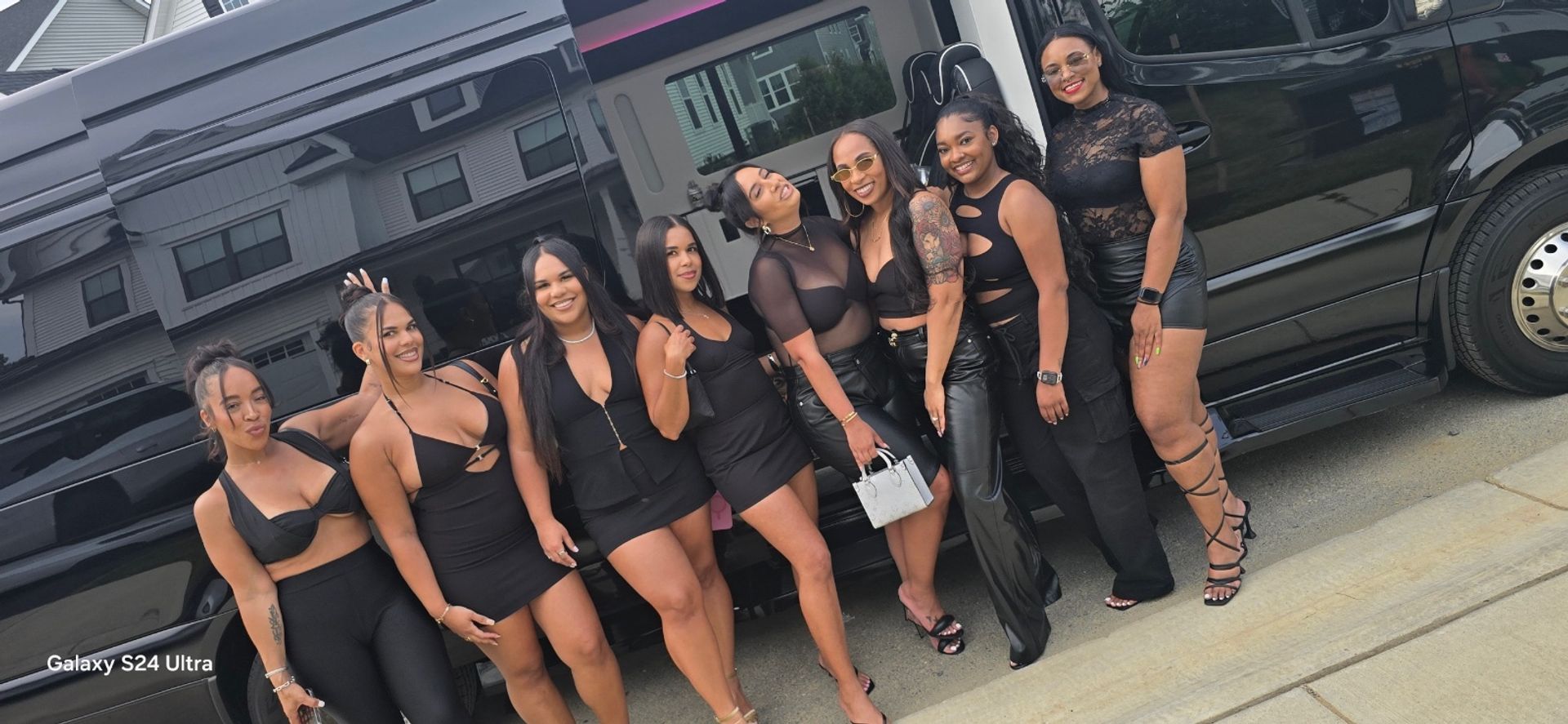 Eight women in coordinated black party outfits smiling and posing beside a glossy black party van on a suburban street, wearing heels, sunglasses and statement accessories for a night out.