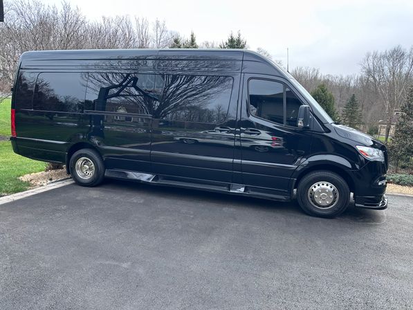 Sleek black high-roof passenger van parked diagonally on a suburban driveway, shiny windows reflecting leafless trees and lawn under an overcast sky