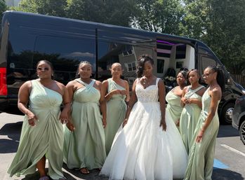Bride in a sparkling white ball gown with six bridesmaids in sage green dresses posing outside beside a black shuttle van on a sunny day