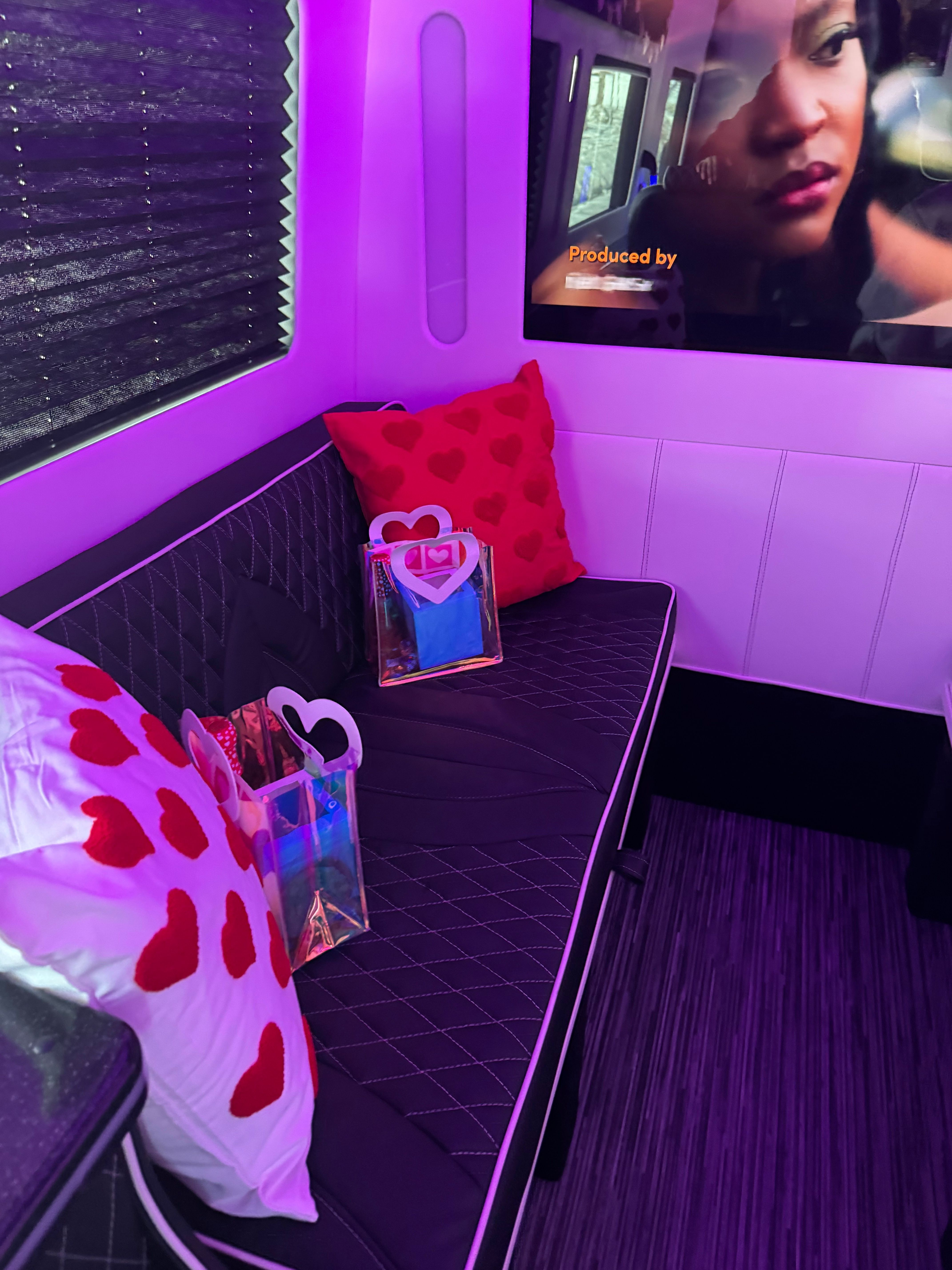 Purple LED-lit party bus lounge interior with a black quilted bench, red heart-pattern pillows, holographic gift bags on the seat, and a wall-mounted TV showing a close-up.