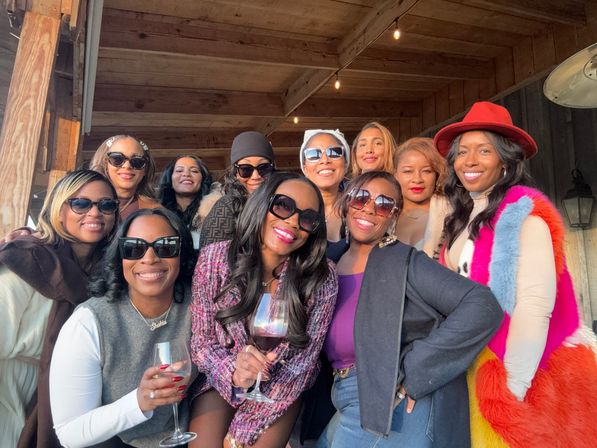 Group selfie of smiling women on a wooden outdoor patio under string lights, wearing winter coats, sunglasses and a red hat, with a couple holding wine glasses for a festive brunch or wine‑tasting vibe.