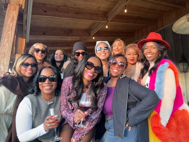Group selfie of smiling women on a wooden outdoor patio under string lights, wearing winter coats, sunglasses and a red hat, with a couple holding wine glasses for a festive brunch or wine‑tasting vibe.