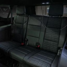 Sleek black leather rear car bench seat with quilted upholstery, three headrests, seatbelts and red buckle latches in a dimly lit vehicle interior.