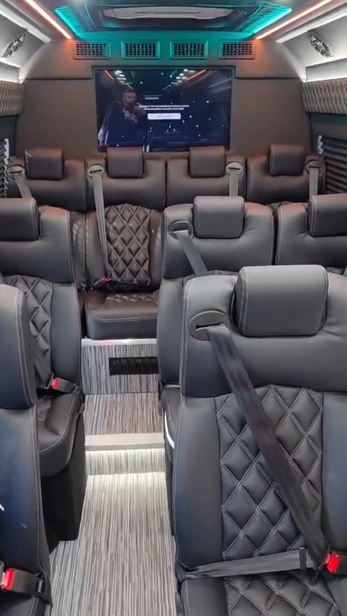 Sleek luxury shuttle van interior with quilted black leather captain seats, visible seatbelts, central aisle and step-up striped flooring, teal and orange LED ambient lighting and a rear flat-screen TV.