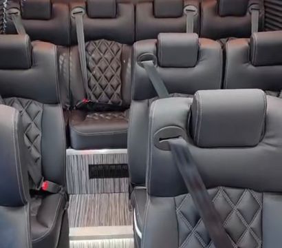 Sleek luxury shuttle van interior with quilted black leather captain seats, visible seatbelts, central aisle and step-up striped flooring, teal and orange LED ambient lighting and a rear flat-screen TV.