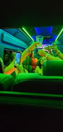 Friends toasting with drinks and a smartphone camera inside a neon-lit party bus with green and blue LED ceiling lights.