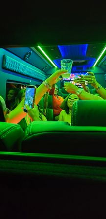 Friends toasting with drinks and a smartphone camera inside a neon-lit party bus with green and blue LED ceiling lights.