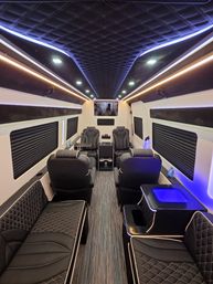 Luxurious passenger van interior with black quilted leather bench and reclining captain chairs, diamond-stitched ceiling, blue and warm LED accent lighting, center aisle and console with cup holders for a sleek VIP shuttle vibe.