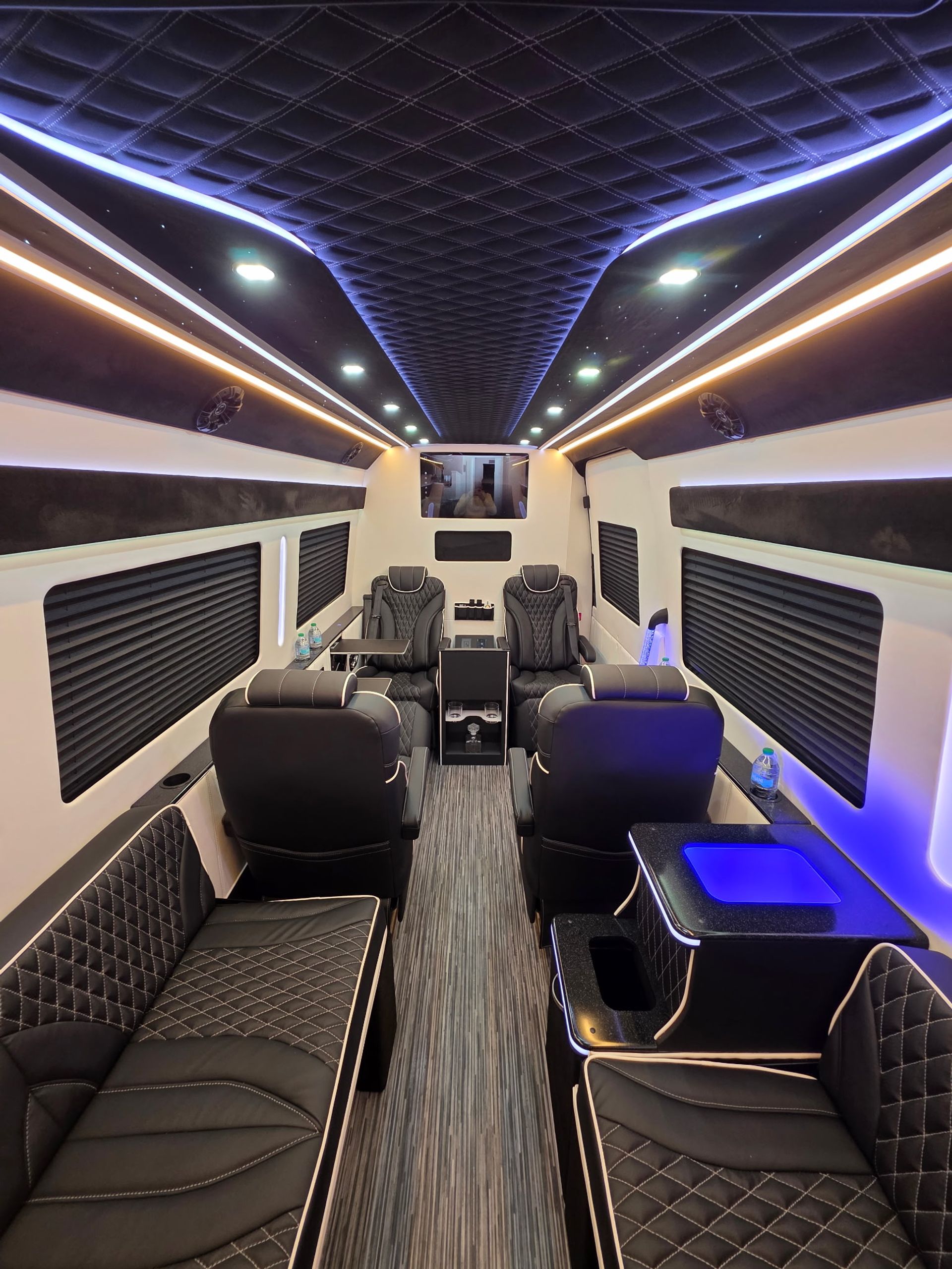 Luxurious passenger van interior with black quilted leather bench and reclining captain chairs, diamond-stitched ceiling, blue and warm LED accent lighting, center aisle and console with cup holders for a sleek VIP shuttle vibe.