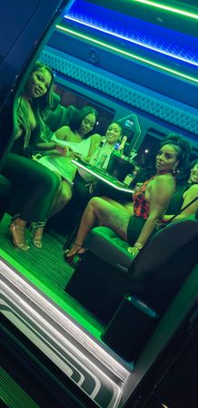 Group of women in a neon-lit luxury party van, seated around a table with bottles and an ice bucket under blue-green LED lights for a night out