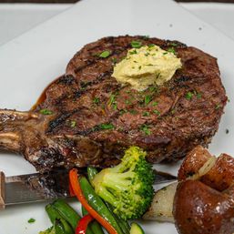 Grilled bone-in ribeye steak topped with melting herb butter, served with roasted baby potatoes and sautéed broccoli, green beans and red pepper on a white plate