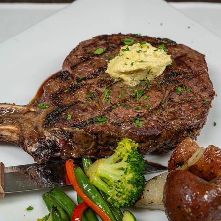 Grilled bone-in ribeye steak topped with melting herb butter, served with roasted baby potatoes and sautéed broccoli, green beans and red pepper on a white plate