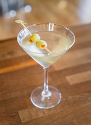 Chilled martini cocktail in a clear glass garnished with two pimento-stuffed green olives on a wooden bar top.