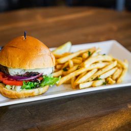 Juicy cheeseburger on a brioche bun with lettuce, tomato and red onion, served with golden French fries and a pickle wedge on a white rectangular plate on a wooden table