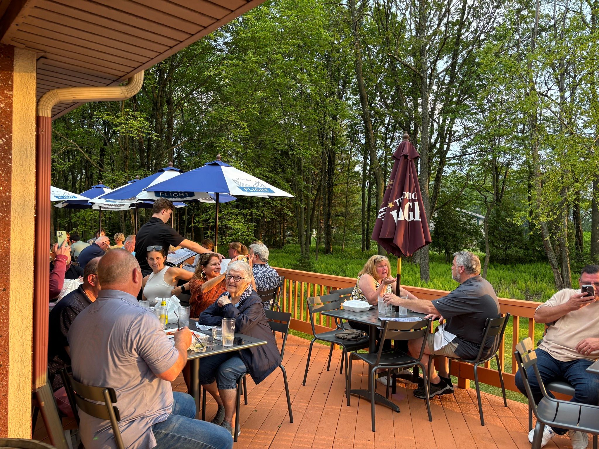 Busy outdoor dining patio on a wooden deck with groups of people at metal tables under blue and brown umbrellas, a server taking orders, and dense green woods beyond at sunset.