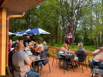 Busy outdoor dining patio on a wooden deck with groups of people at metal tables under blue and brown umbrellas, a server taking orders, and dense green woods beyond at sunset.