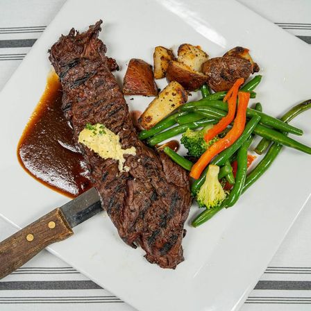 Grilled flank steak topped with herb butter and a rich pan sauce, served with roasted red potatoes and sautéed green beans, carrots and broccoli on a white square plate with a wooden-handled steak knife.