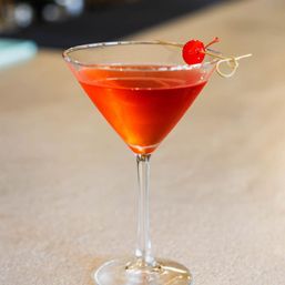 Vibrant red cocktail in a martini glass topped with a maraschino cherry on a cocktail pick, set on a light bar counter.