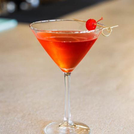 Vibrant red cocktail in a martini glass topped with a maraschino cherry on a cocktail pick, set on a light bar counter.